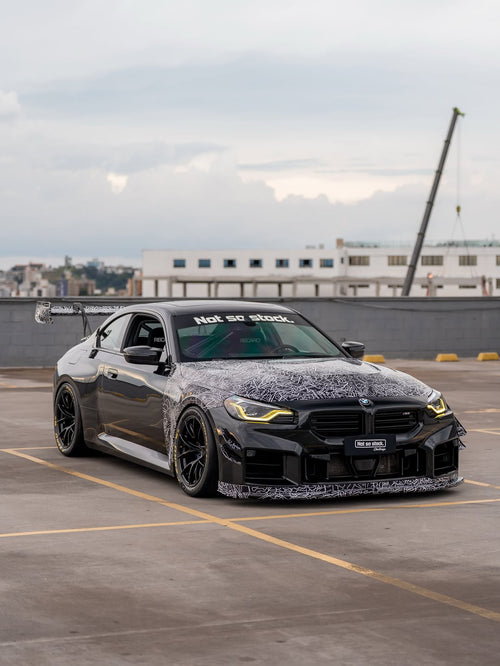 Gray BMW M2 featuring a REVOZPORT body kit with front lip, rear wing, canards, vented fenders, and hood, parked in an open rooftop lot.