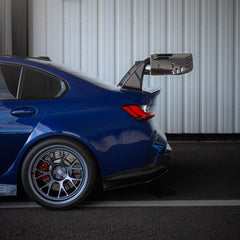 Blue BMW G80 M3 with aggressive carbon fiber aero kit, front splitter, vented fenders, and side skirts parked beside a professional race track during sunset.