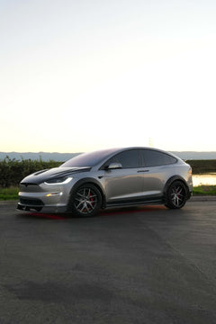 Tesla Model X featuring an aggressive REVOZPORT carbon fiber body kit with front lip, vented hood, wide fender extensions, side skirts, and illuminated red underglow lighting at dusk.