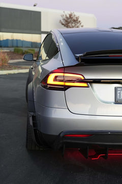 Tesla Model X featuring an aggressive REVOZPORT carbon fiber body kit with front lip, vented hood, wide fender extensions, side skirts, and illuminated red underglow lighting at dusk.