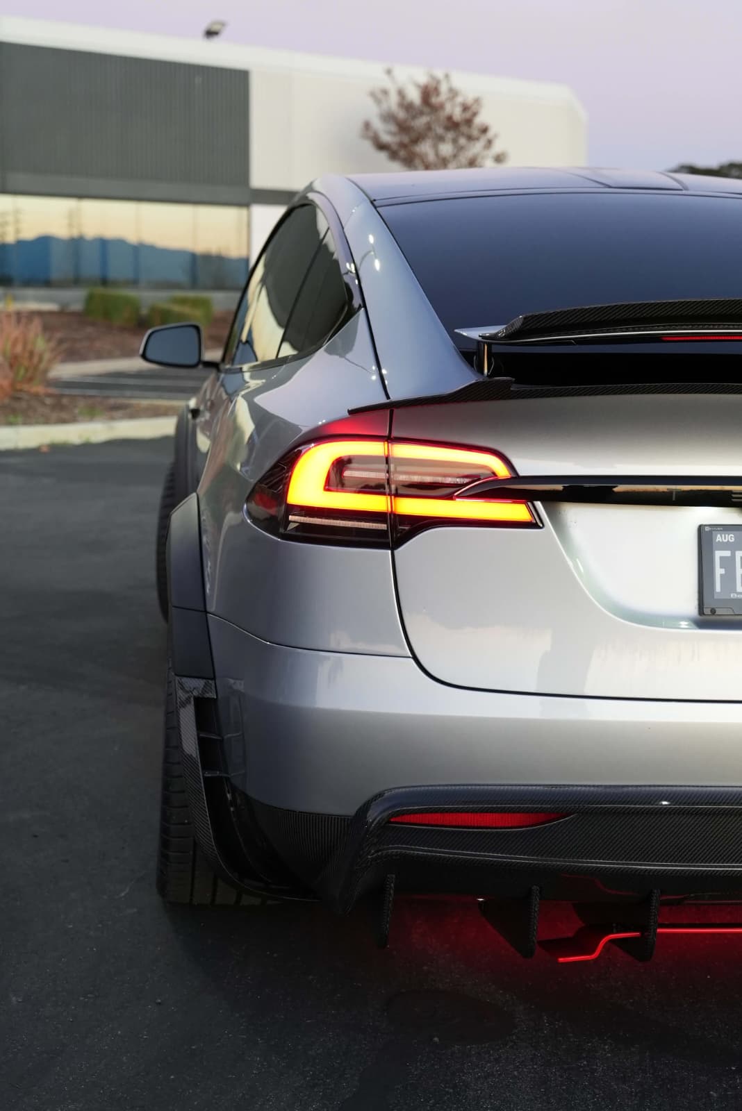 Tesla Model X featuring an aggressive REVOZPORT carbon fiber body kit with front lip, vented hood, wide fender extensions, side skirts, and illuminated red underglow lighting at dusk.