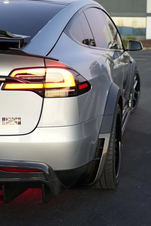 Tesla Model X featuring an aggressive REVOZPORT carbon fiber body kit with front lip, vented hood, wide fender extensions, side skirts, and illuminated red underglow lighting at dusk.