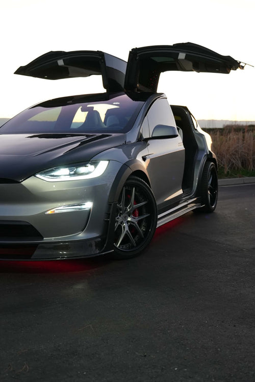 Tesla Model X featuring an aggressive REVOZPORT carbon fiber body kit with front lip, vented hood, wide fender extensions, side skirts, and illuminated red underglow lighting at dusk.