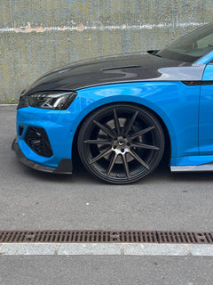 Blue Audi RS5 Sportback featuring a REVOZPORT carbon fiber body kit with vented hood, front lip, side skirts, and lowered stance parked outside a modern industrial building at sunset.
