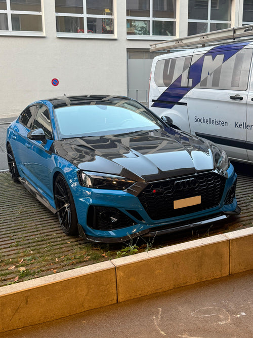 Blue Audi RS5 Sportback featuring a REVOZPORT carbon fiber body kit with vented hood, front lip, side skirts, and lowered stance parked outside a modern industrial building at sunset.