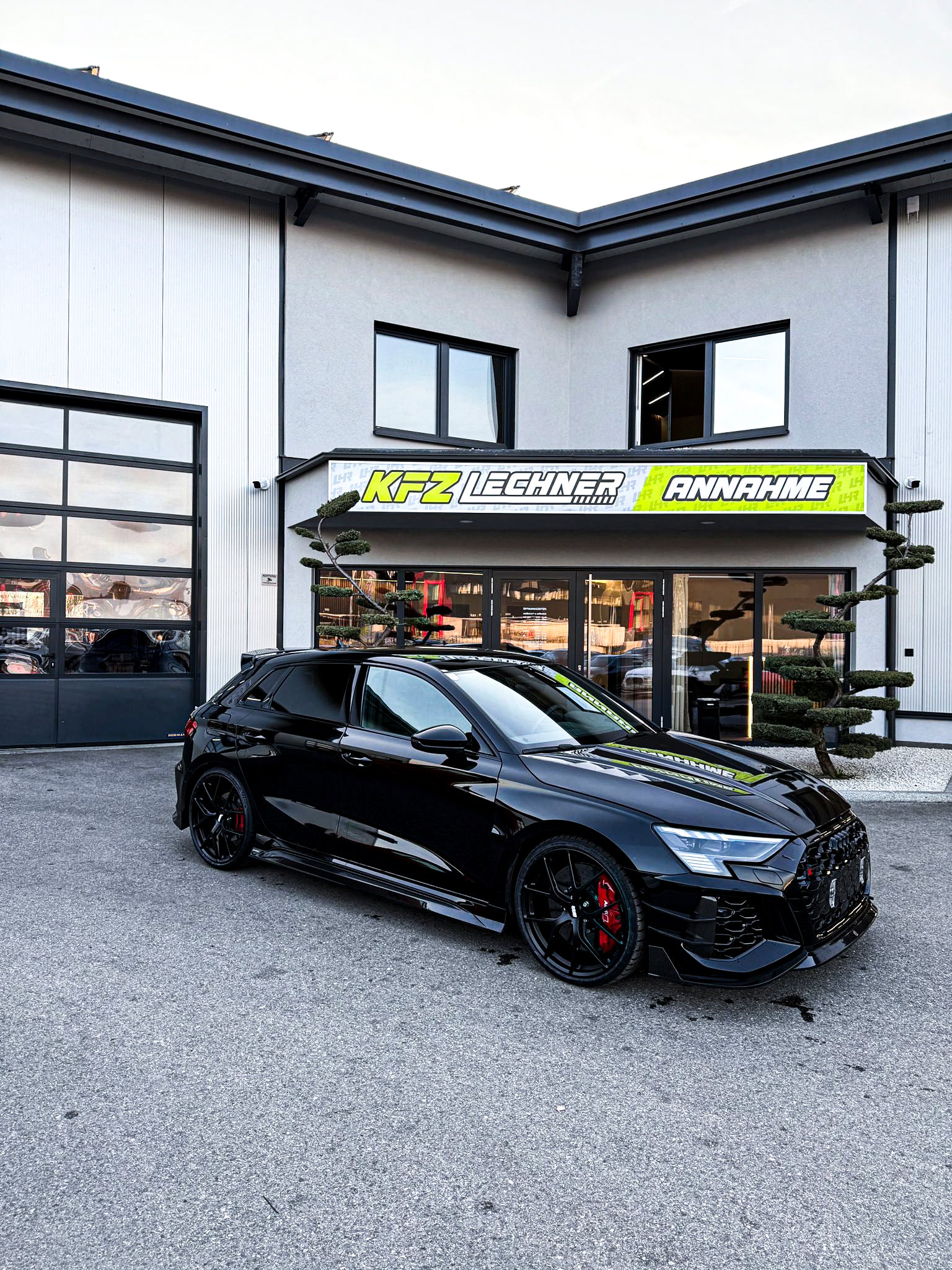 Black Audi RS3 Sportback featuring a REVOZPORT carbon fiber body kit with front lip and side skirts parked on a wet outdoor lot.