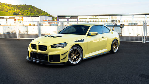 Front three-quarter view of a BMW featuring aggressive carbon fiber aero, vented hood, bronze forged wheels, and oversized rear wing styling.