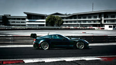 Black Chevrolet Corvette race car parked on a wet industrial track surface, featuring an aggressive wide-body kit, large rear wing, carbon fiber aerodynamic components, and lightweight performance wheels with red brake calipers. The moody urban backdrop and reflective pavement emphasize the car’s high-performance motorsport styling.
