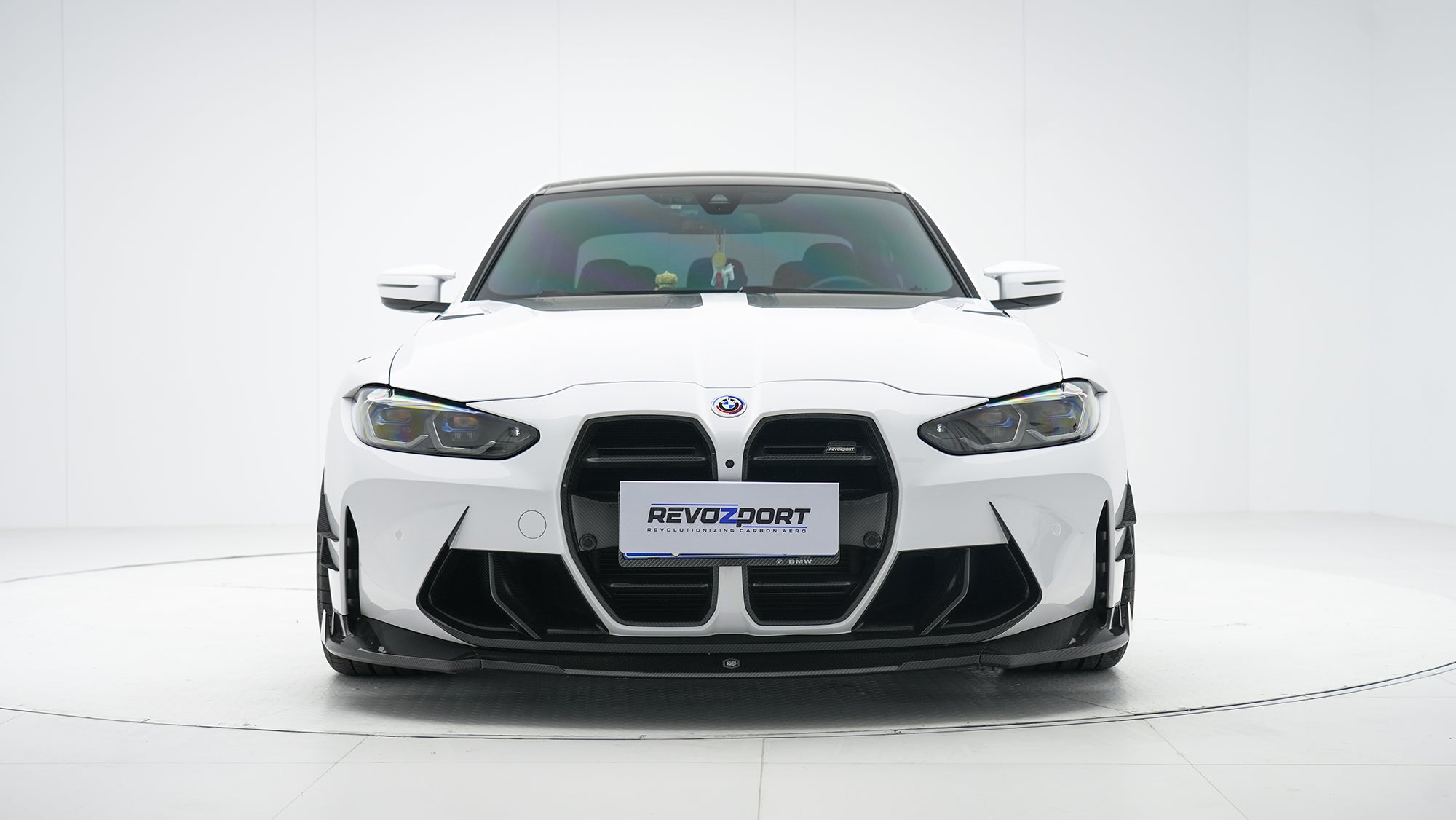 White BMW G80 M3 featuring a REVOZPORT carbon fiber body kit with front lip, side skirts, canards, vented fenders, and yellow DRL headlights displayed in a clean studio setting.