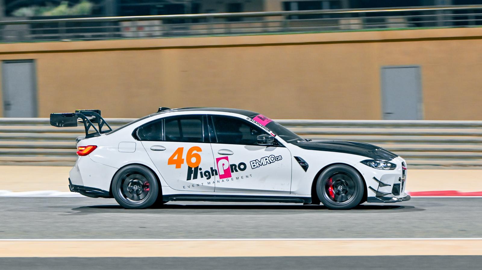 White BMW G80 M3 track car with REVOZPORT carbon fiber aero kit, front canards, rear wing, and racing decals driving on a professional race circuit.