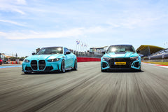 Two turquoise performance cars with carbon fiber aero kits driving side by side on a racetrack under bright sunlight.