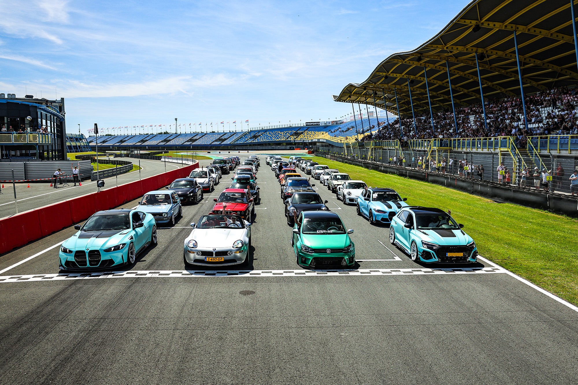 Multiple modified performance cars lined up on a racetrack grid, showcasing carbon fiber aero styling at a motorsport event.