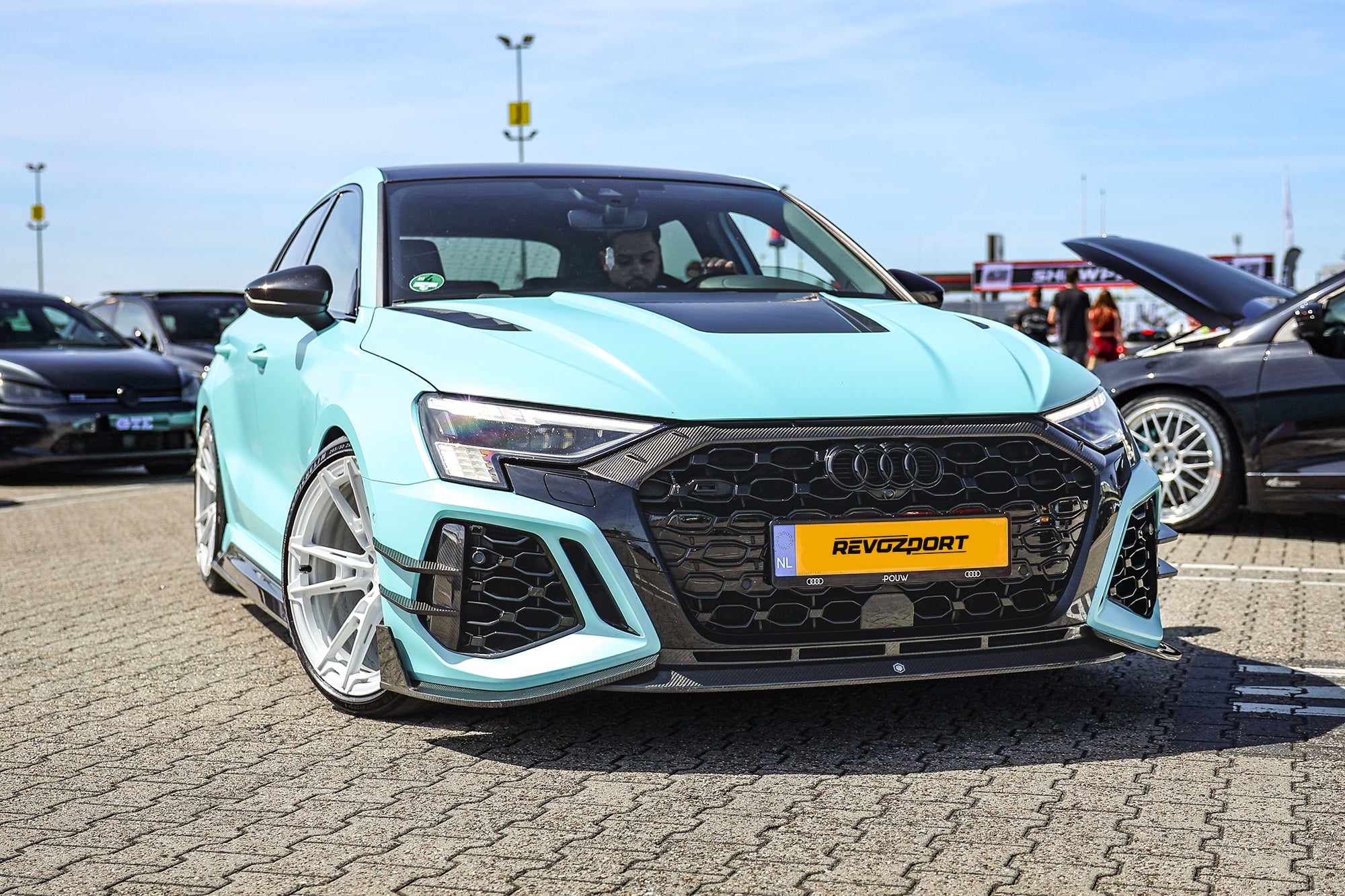 Turquoise Audi with carbon fiber body kit and white wheels displayed at an outdoor car event, highlighting aggressive front aero styling.