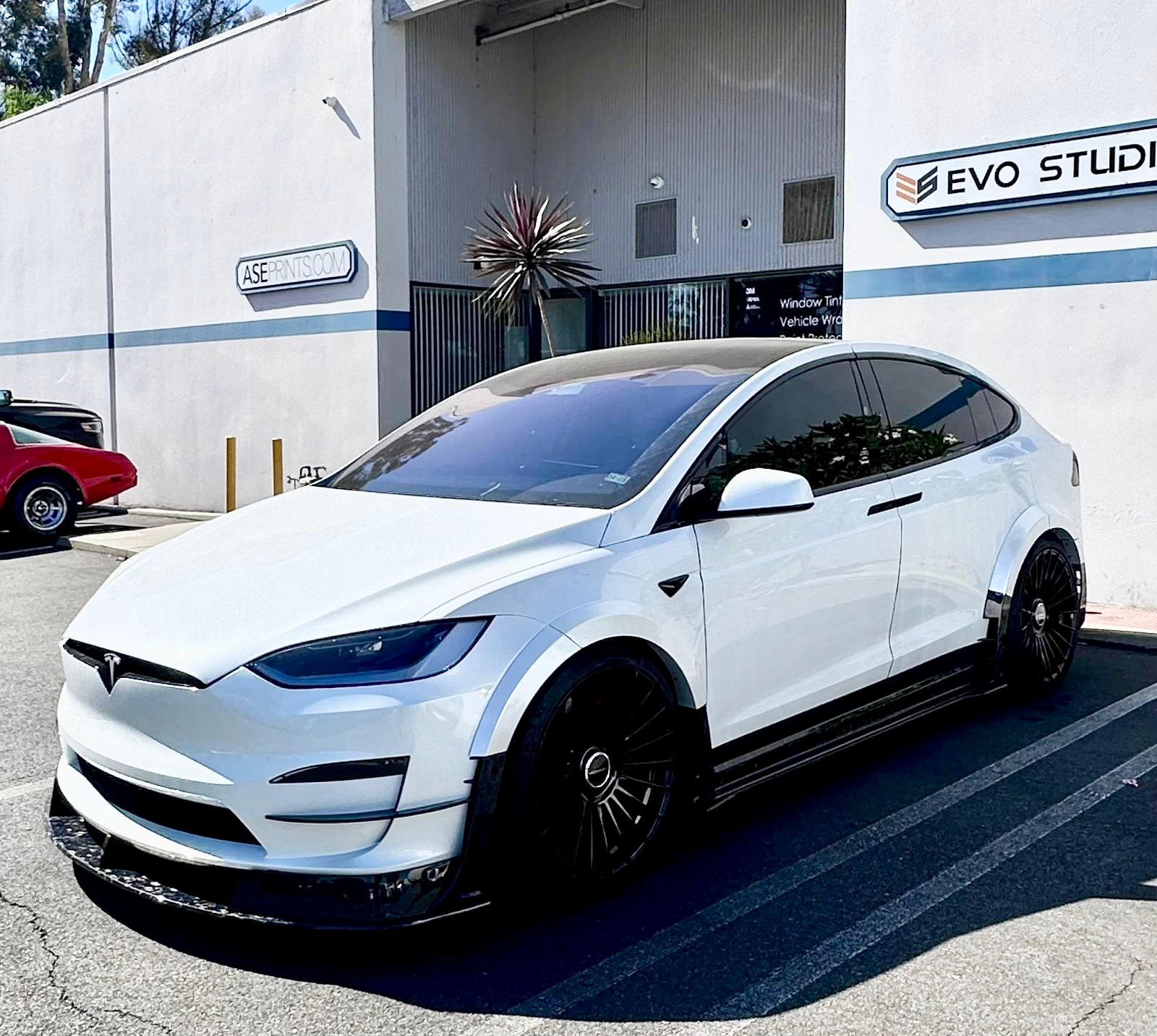 White Tesla Model X featuring an aggressive REVOZPORT body kit with carbon fiber front lip, side skirts, wide fender extensions, and lowered stance parked outside a detailing studio.