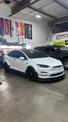 White Tesla Model X featuring an aggressive REVOZPORT body kit with carbon fiber front lip, side skirts, wide fender extensions, and lowered stance parked outside a detailing studio.