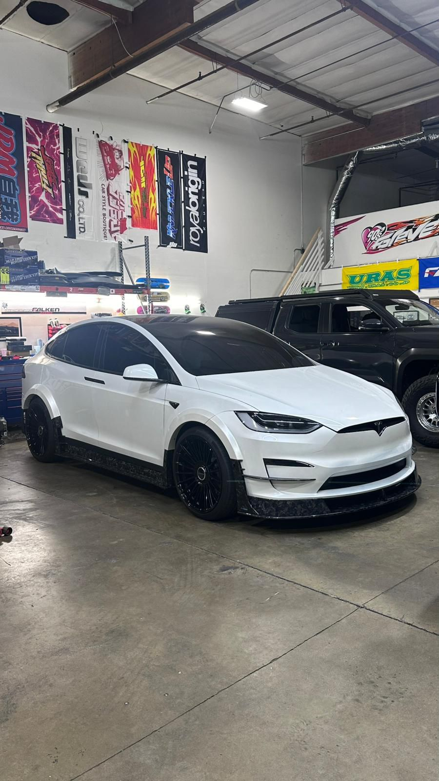 White Tesla Model X featuring an aggressive REVOZPORT body kit with carbon fiber front lip, side skirts, wide fender extensions, and lowered stance parked outside a detailing studio.
