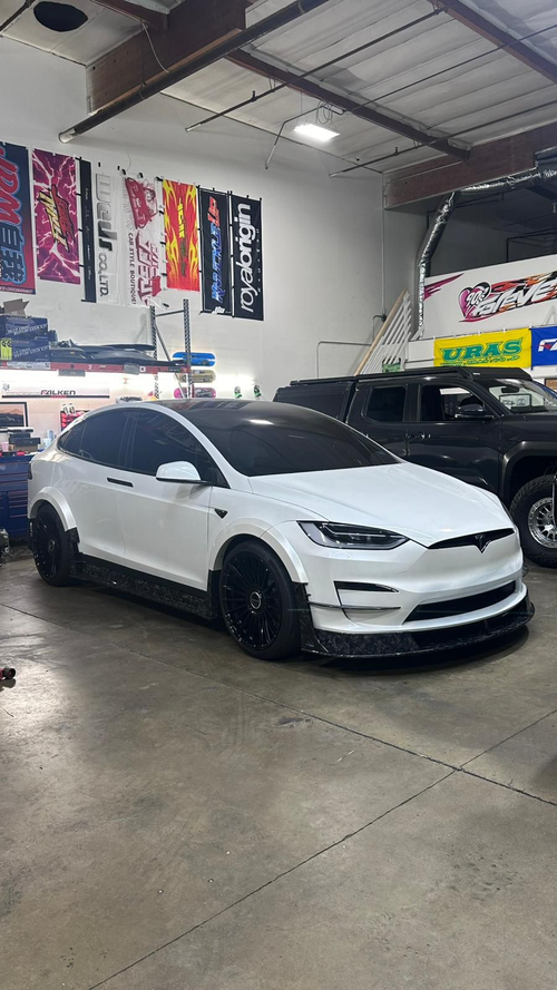 White Tesla Model X featuring an aggressive REVOZPORT body kit with carbon fiber front lip, side skirts, wide fender extensions, and lowered stance parked outside a detailing studio.
